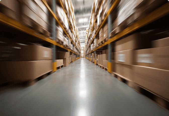A wide warehouse aisle lined with tall shelving units stacked with brown cardboard boxes, shot with a motion blur effect.