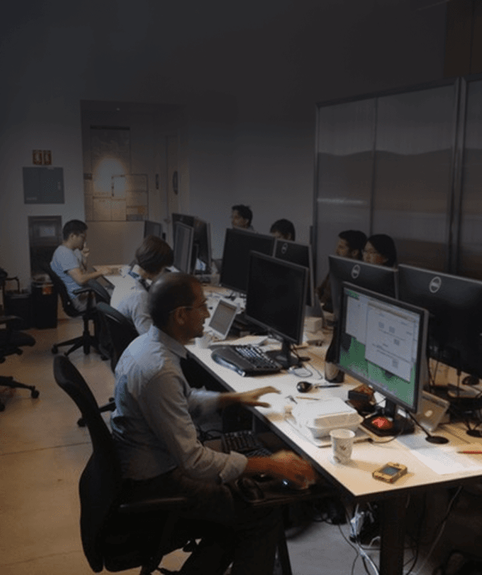 Several office workers sit at desks with computers in a dimly lit open workspace, focused on their screens.