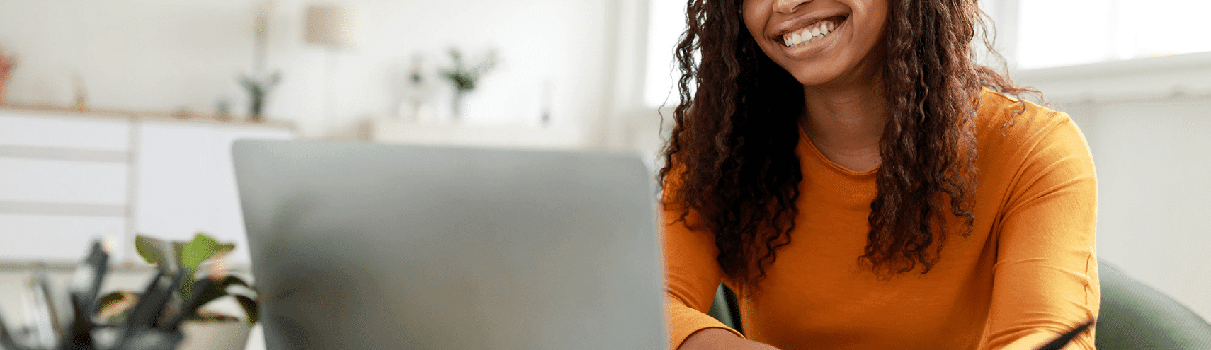 A young college student smiling while wearing a bright orange sweater as she sits down in front of her laptop