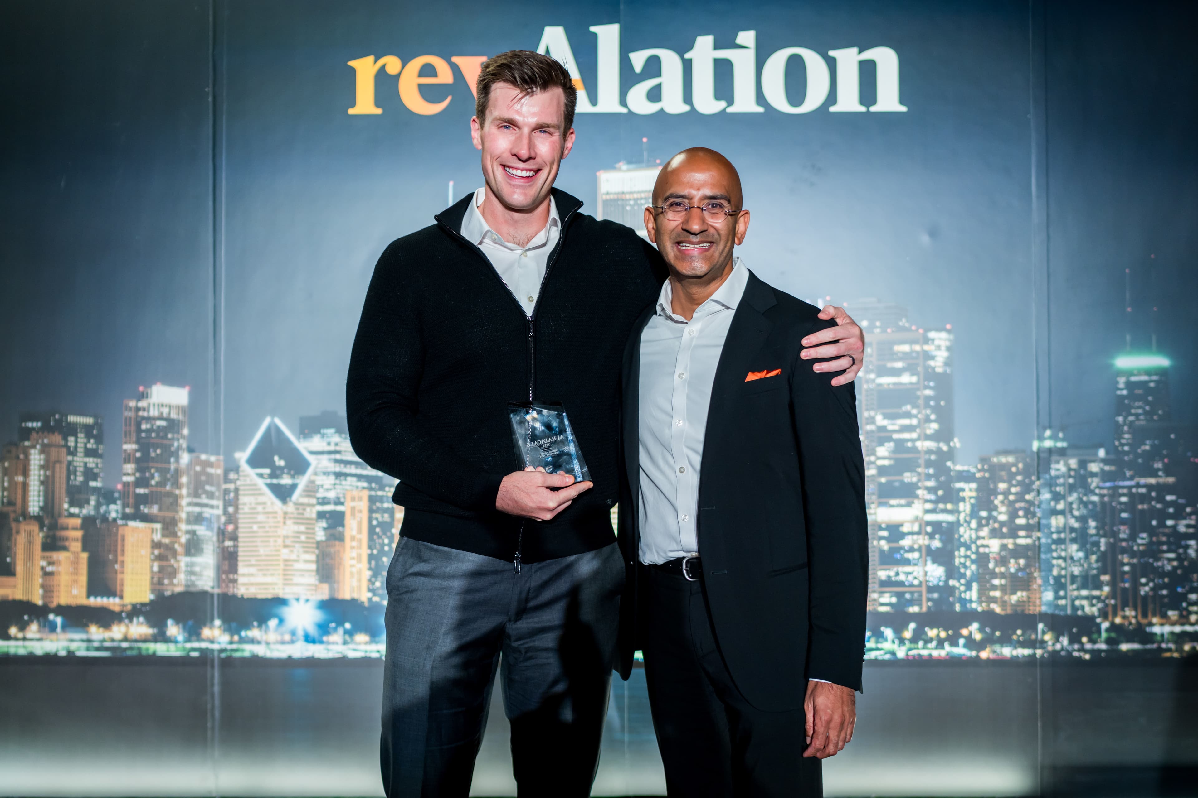 Two smiling men pose together at a RevAlation event, one holding a crystal award, with a city skyline backdrop.