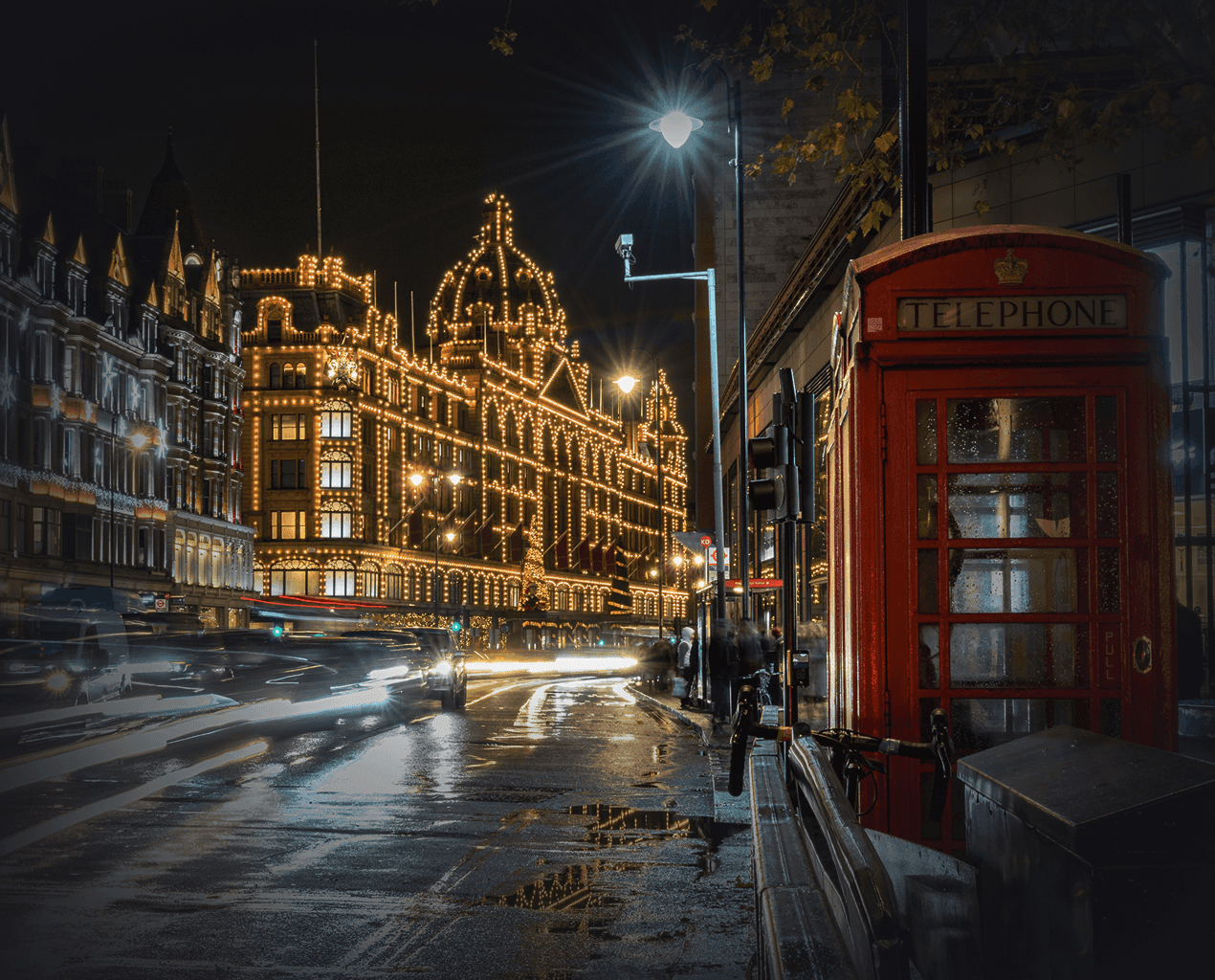 A red London telephone box at night with Harrods department store lit up with golden lights in the background.