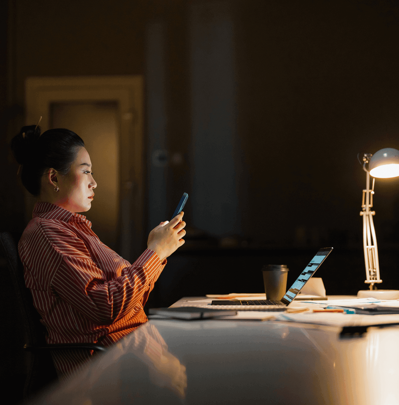 Woman in a red striped shirt working late at a dimly lit desk, using a smartphone with a laptop and lamp nearby.