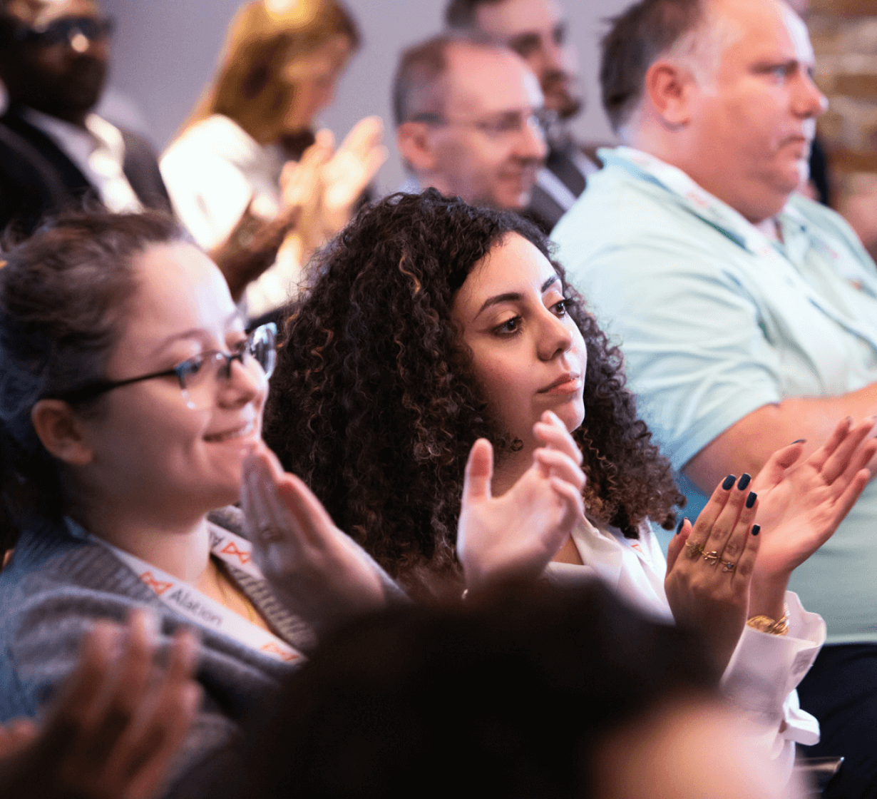 Diverse audience members applauding at a conference, with focus on a woman with curly hair in the foreground.
