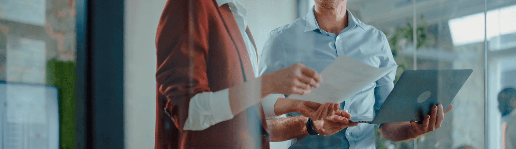 A male coworker and a female coworker in a work setting discussing data while holding their laptops and data sheets