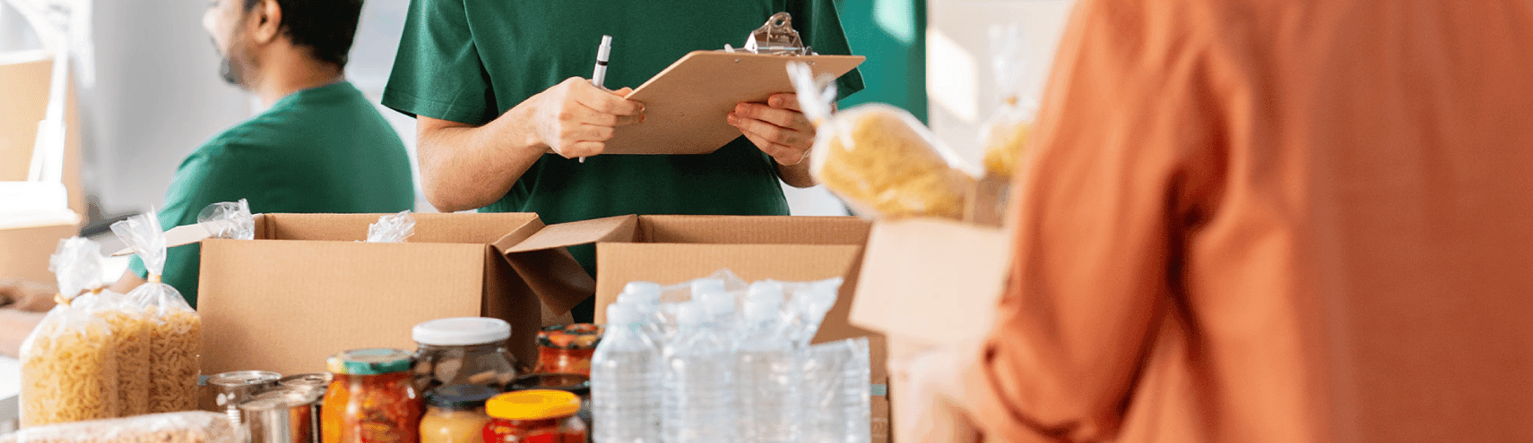 A volunteer holding a clipboard as someone brings in a box of food donations to a table