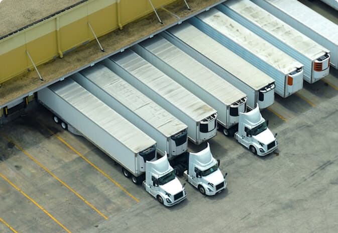 Aerial view of multiple white semi-trucks with trailers docked at a yellow warehouse loading bay.