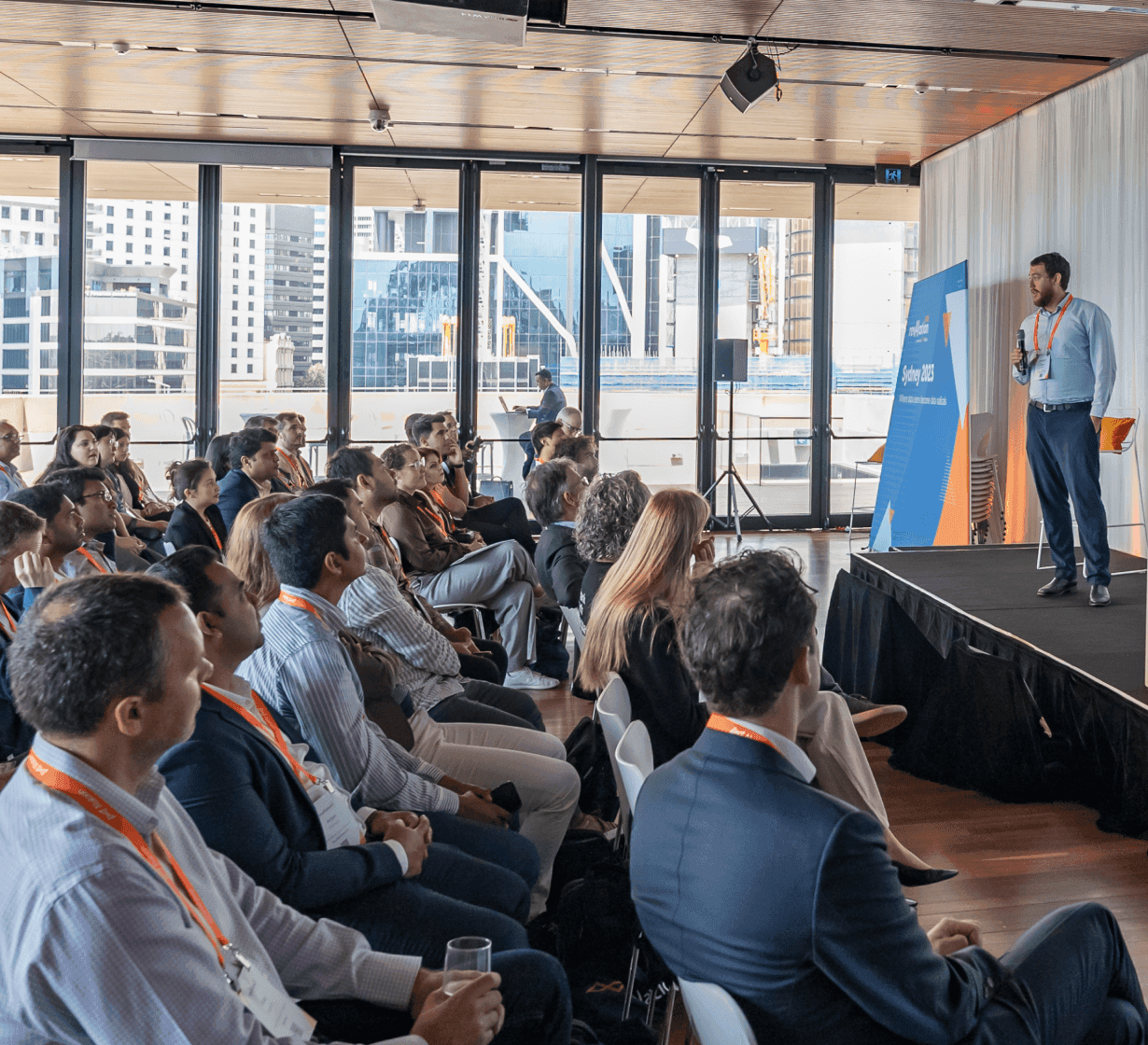 Speaker presenting at a business conference with attendees in a modern venue featuring large windows overlooking city buildings.