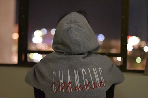 A person in a stylish grey hoodie with the empowering message 'Changing Mindsets' on the back, gazing contemplatively at the city lights.