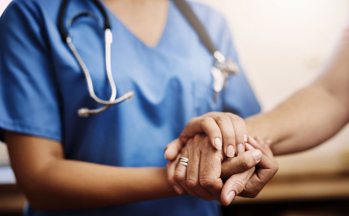 Healthcare professional in blue scrubs with stethoscope comforting a patient by holding their hands.