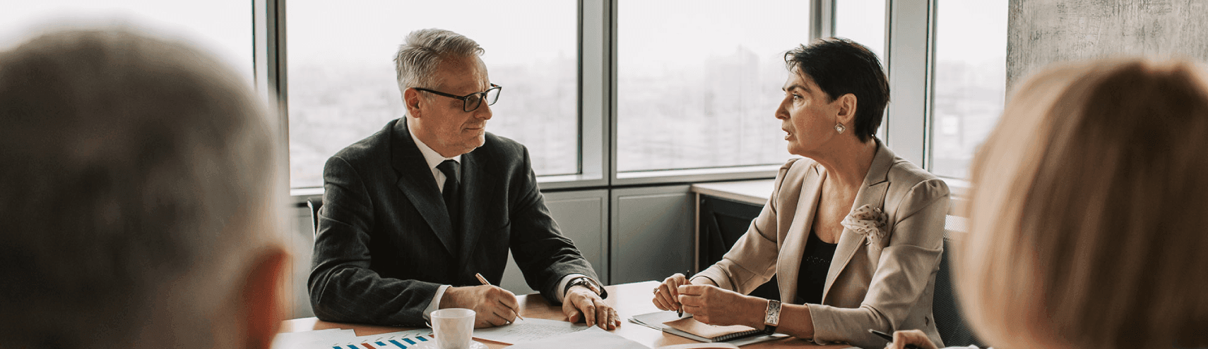 Alation Blog Image: An elderly man and woman sitting across a table, having a discussion about business. The man is holding a pen and a paper, while the woman looks attentive towards him.