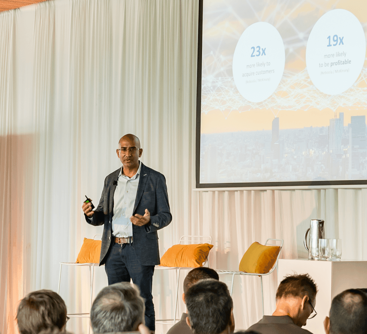 Speaker presenting business statistics showing 23x customer acquisition and 19x profitability metrics to audience