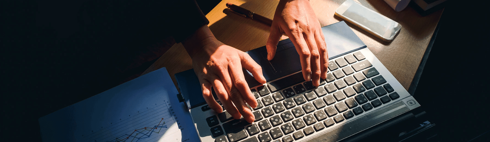 Birds-eye view of a man's hands over his laptop on top of a desk with notebooks and loose leaf paper with graphs on them