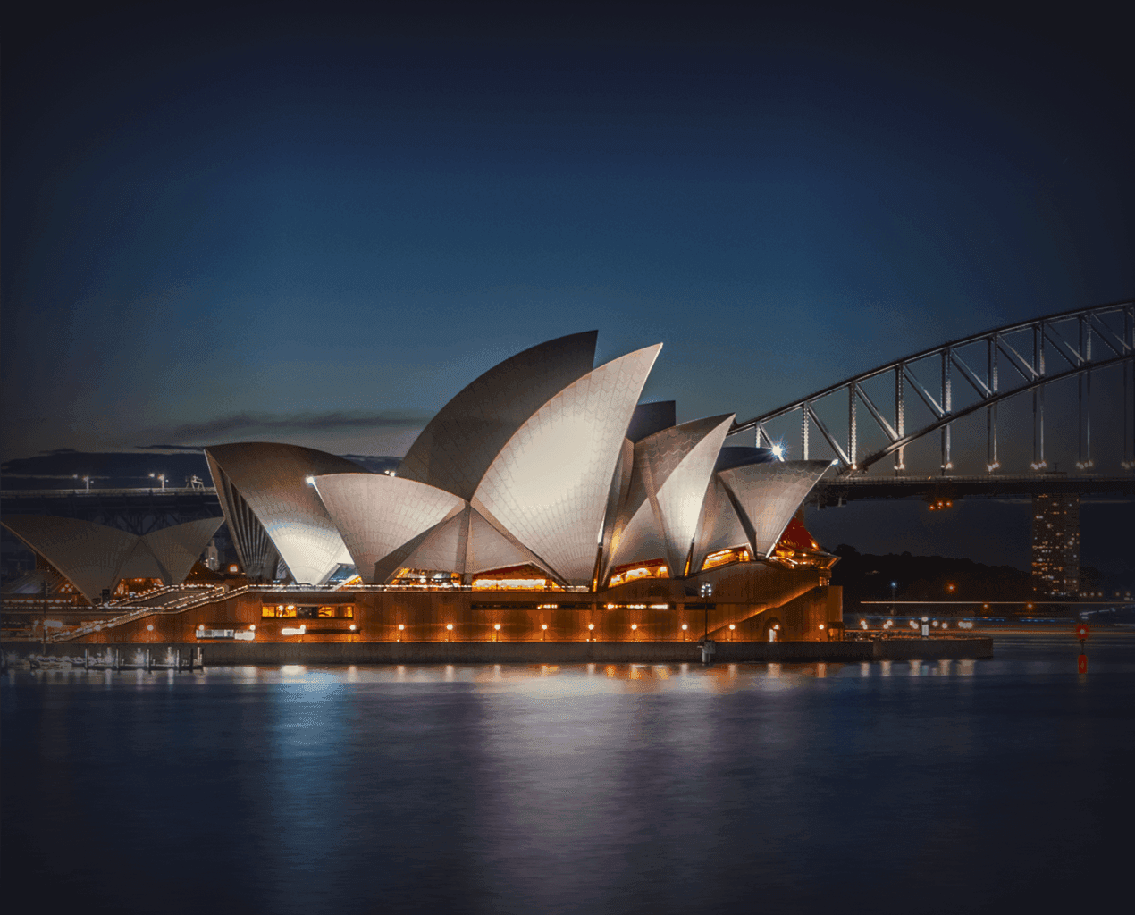 Sydney Opera House illuminated at night with the Harbour Bridge in the background, reflecting warm lights on the dark harbor water.