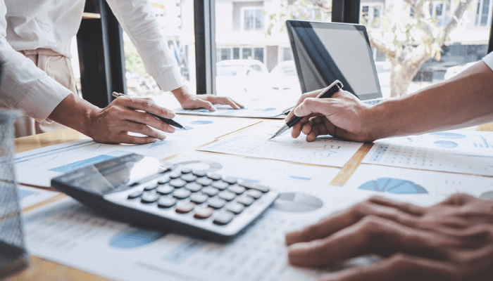 Alation blog image: A bustling desk with hands diligently jotting down notes on various graphs placed on loose-leaf papers. A calculator and laptop accompany the scene, showcasing a dynamic work environment.