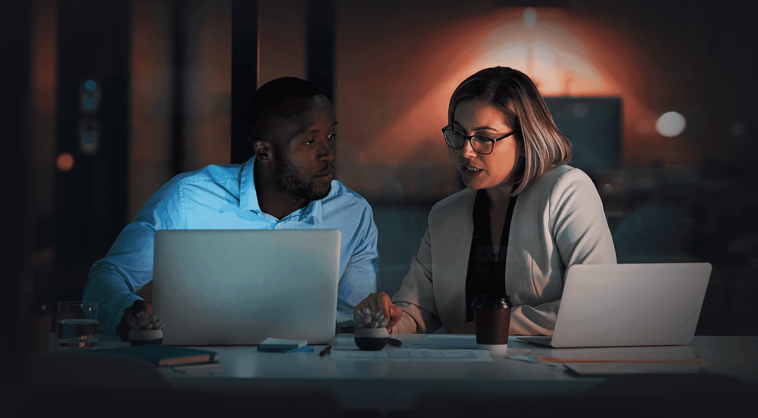 Two colleagues collaborating at a dimly lit office desk at night, each with a laptop, engaged in focused discussion.