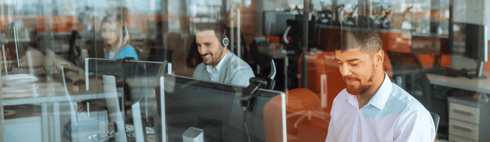 A reflective window view of a modern office space with three coworkers working together on their desktop computers