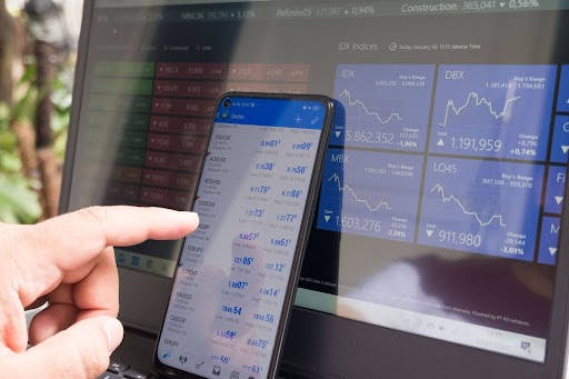 A hand points to stock prices on a phone with a laptop showing market graphs in the background.