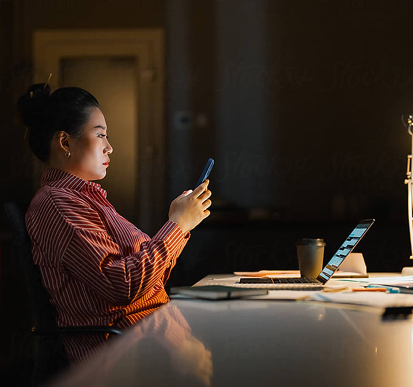 Woman in a red striped shirt working late at a dimly lit desk, holding a smartphone with a laptop open nearby.