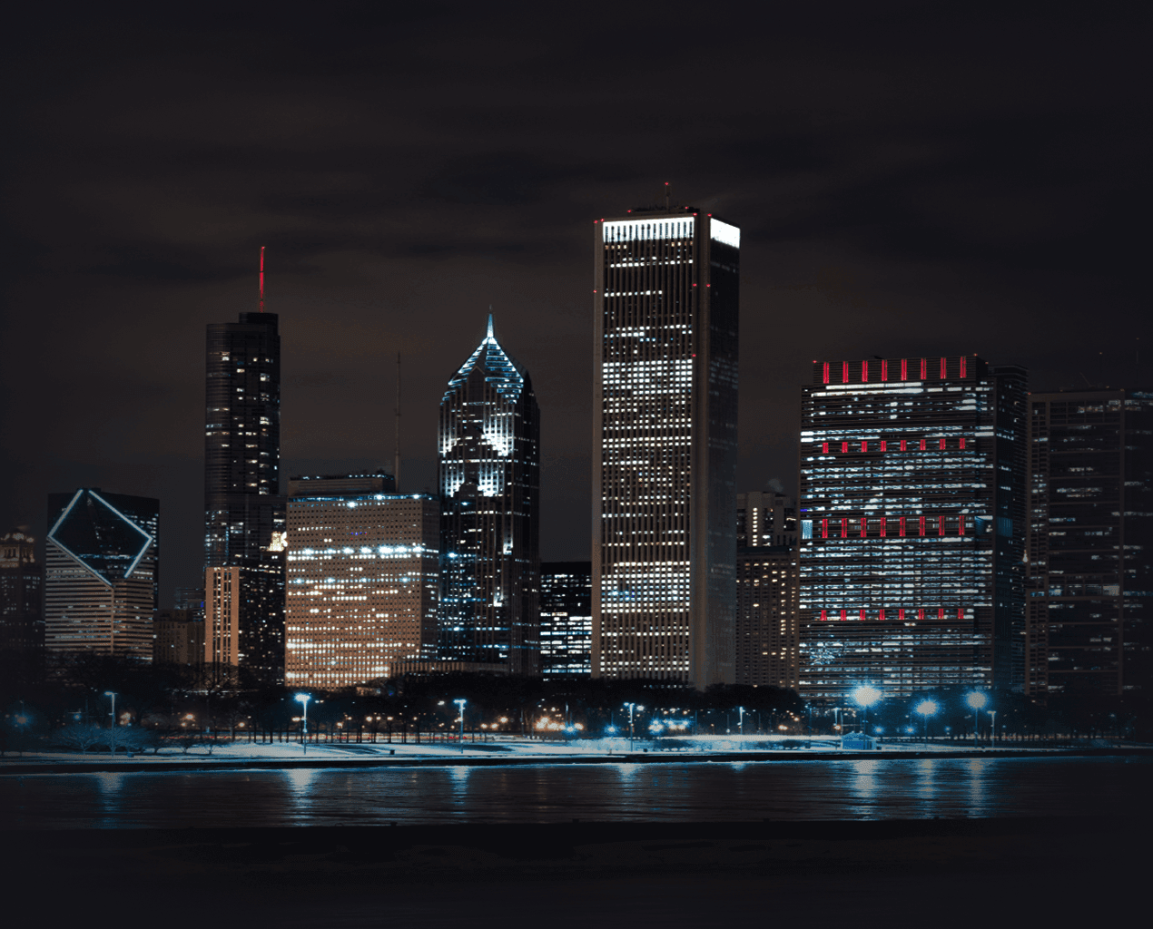 Chicago skyline at night with illuminated skyscrapers reflecting on Lake Michigan under a dark sky.